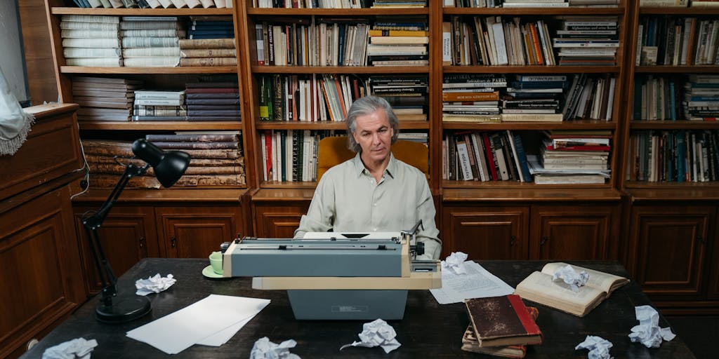 Cinematic shot of a lone writer at a vintage wooden desk late at night. A continuous scroll of paper (one long sheet) unspools from a fountain pen across the desk, down onto the floor and out of frame, filled with dense typed paragraphs, margin notes and colorful sticky flags marking sections. Warm desk lamp light creates deep shadows; a coffee cup, glasses and an hourglass sit nearby to emphasize time investment. Framing: low angle to emphasize the length of the document receding into darkness.