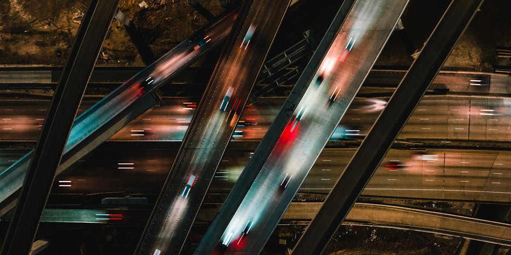 Aerial/drone shot of a normally busy multi-lane highway at night where car light trails suddenly stop at one interchange, creating an eerie empty gap; creative post-processing overlays a translucent descending traffic-graph line and map pins above the road to visually link physical traffic loss with data analysis.