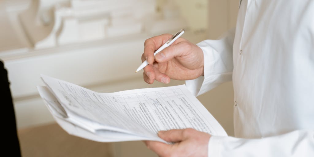 Playful editorial image of an SEO 'doctor' in a white coat and stethoscope diagnosing a giant website icon on a desk. The doctor holds a clipboard showing a checklist (site audit, backlinks, DR score) and points to a highlighted 'DR: 42' label on the site, evoking the idea of a site health check focused on Domain Rating.