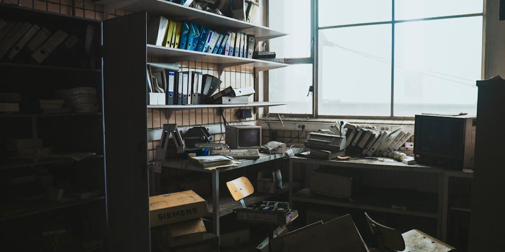 A dusty attic filled with old cardboard boxes and cobwebs; one open box reveals manila folders labeled “Posts,” “Pages,” and “Drafts.” In the foreground sits a vintage laptop showing the WordPress dashboard with an untouched post highlighted. Dust motes in the sunlight and a sense of long-forgotten files visually communicate neglected or orphaned content.
